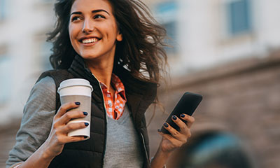 Women walking in public holding her phone and cup of coffee.